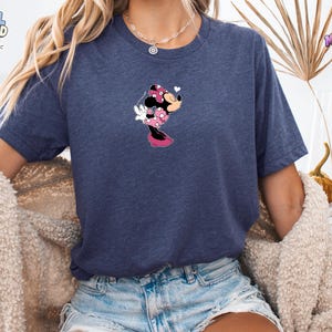 May include: A heather navy blue short-sleeve t-shirt featuring a small graphic of Minnie Mouse wearing a pink polka-dot dress and bow, blowing a kiss. The character is positioned centrally on the chest of the shirt, which has a crew neckline.