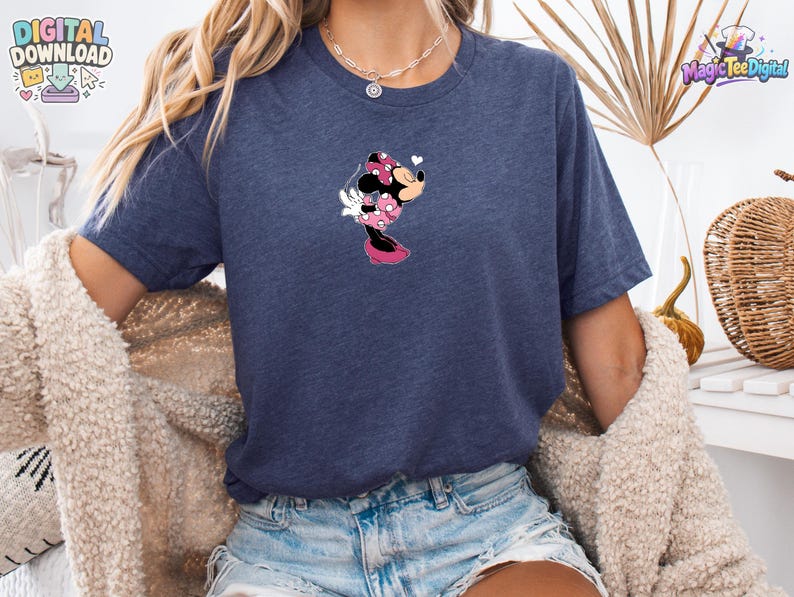 May include: A heather navy blue short-sleeve t-shirt featuring a small graphic of Minnie Mouse wearing a pink polka-dot dress and bow, blowing a kiss. The character is positioned centrally on the chest of the shirt, which has a crew neckline.