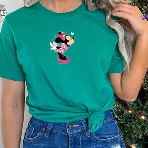 May include: A bright green short-sleeved t-shirt featuring a centered graphic of Minnie Mouse wearing a pink polka-dot dress and bow, blowing a kiss with a small white heart above her head. The shirt is paired with blue denim jeans.