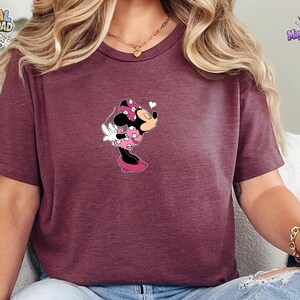 May include: A maroon short-sleeved t-shirt featuring a graphic of Minnie Mouse wearing a pink polka-dot dress and bow, leaning forward with a small white heart icon above her head. The shirt has a crew neckline and a heathered fabric texture.