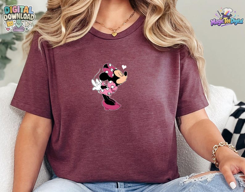 May include: A maroon short-sleeved t-shirt featuring a graphic of Minnie Mouse wearing a pink polka-dot dress and bow, leaning forward with a small white heart icon above her head. The shirt has a crew neckline and a heathered fabric texture.