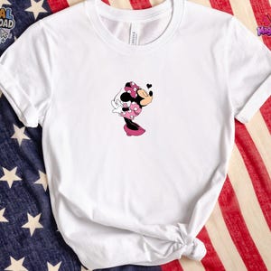 May include: A white short-sleeved crew neck t-shirt featuring a small graphic of Minnie Mouse in a pink polka-dot dress and bow, blowing a kiss with a small black heart icon above her head. The shirt has a simple, clean design on the front.