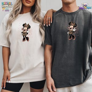 May include: A cream-coloured t-shirt and a charcoal grey t-shirt, each featuring a graphic of a cartoon mouse character wearing a leopard print dress and pink sunglasses, surrounded by small pink glitter-like specks and accents.