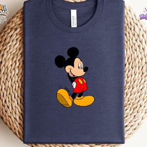 Peut inclure: Un t-shirt bleu marine &agrave; col rond arborant une impression graphique de Mickey Mouse. Le personnage est repr&eacute;sent&eacute; dans une pose classique avec des oreilles noires, un visage beige, un short rouge et de grandes chaussures jaunes.