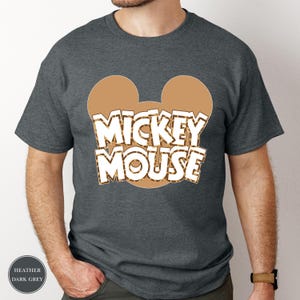Puede incluir: Camiseta gris oscuro jaspeado con un contorno de cabeza de Mickey Mouse marr&oacute;n y un estampado de leopardo. Las palabras "MICKEY MOUSE" son blancas con un contorno marr&oacute;n. La camiseta es de cuello redondo y manga corta.