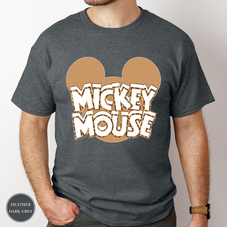 Puede incluir: Camiseta gris oscuro jaspeado con un contorno de cabeza de Mickey Mouse marr&oacute;n y un estampado de leopardo. Las palabras "MICKEY MOUSE" son blancas con un contorno marr&oacute;n. La camiseta es de cuello redondo y manga corta.