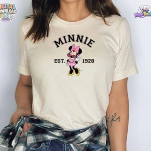 May include: A cream-colored short-sleeve t-shirt featuring a graphic of Minnie Mouse wearing pink sunglasses and a polka dot dress. The text above the character reads MINNIE in black, and the text below reads EST. 1928 in black lettering.
