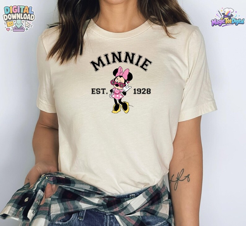 May include: A cream-colored short-sleeve t-shirt featuring a graphic of Minnie Mouse wearing pink sunglasses and a polka dot dress. The text above the character reads MINNIE in black, and the text below reads EST. 1928 in black lettering.