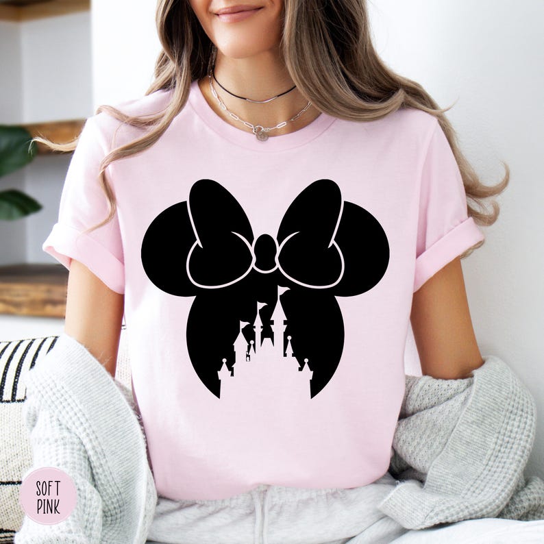 May include: A light pink t-shirt featuring a black silhouette of a bow with a castle design. The words "SOFT PINK" are printed on the bottom left of the shirt. The shirt has short sleeves and a crew neck.