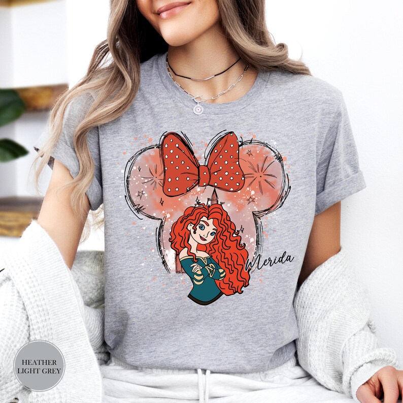 Puede incluir: Camiseta gris claro con una ilustraci&oacute;n de dibujos animados de Merida de Brave. El dise&ntilde;o incluye un lazo rojo con lunares blancos, un personaje de Merida y el nombre "Merida" en escritura. La camiseta es de color gris claro.