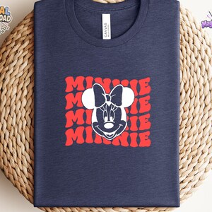 May include: A navy blue t-shirt featuring a white graphic of Minnie Mouse's face centered over a repeating red wavy text pattern that reads MINNIE. The shirt has a crew neckline and a standard fit, showcasing a clean and simple design aesthetic.