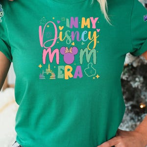 May include: A green short-sleeved t-shirt featuring a colorful graphic print that reads "IN MY Disney MOM ERA" in various pastel colors. The design includes a purple mouse-ear silhouette with a bow, a castle icon, and a hand gesture icon.