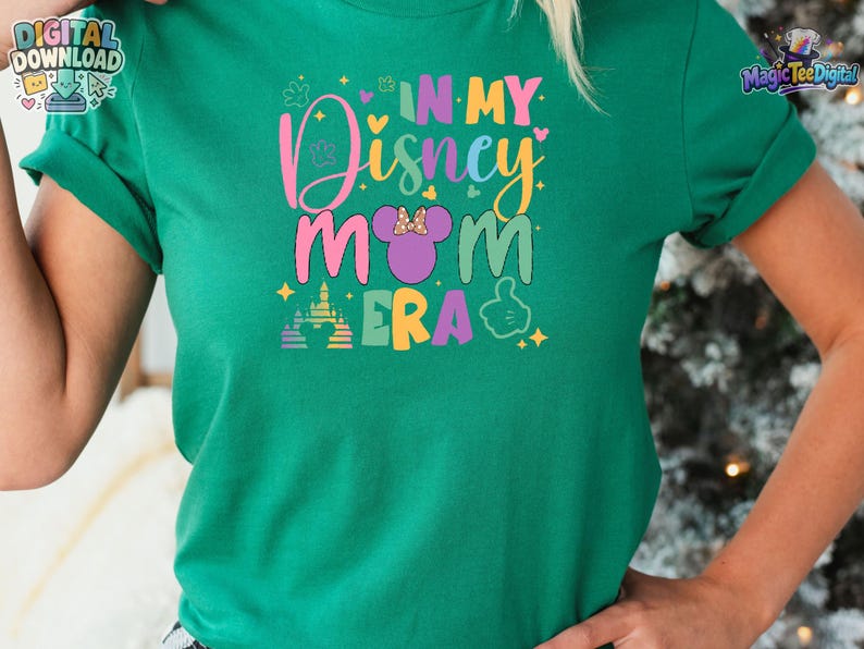 May include: A green short-sleeved t-shirt featuring a colorful graphic print that reads "IN MY Disney MOM ERA" in various pastel colors. The design includes a purple mouse-ear silhouette with a bow, a castle icon, and a hand gesture icon.