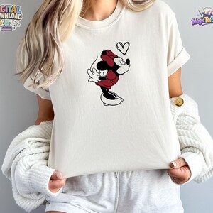 May include: A cream-colored short-sleeve t-shirt featuring a graphic of a cartoon mouse character wearing a red dress and bow, leaning forward with eyes closed, accompanied by a small black outline of a heart floating above the character's head.
