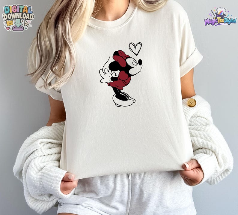May include: A cream-colored short-sleeve t-shirt featuring a graphic of a cartoon mouse character wearing a red dress and bow, leaning forward with eyes closed, accompanied by a small black outline of a heart floating above the character's head.