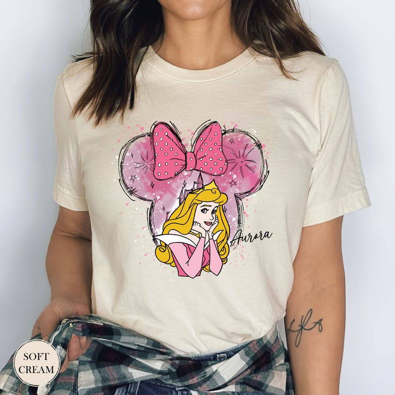 May include: Soft cream t-shirt with a graphic of Princess Aurora, a pink bow, and a castle. The design includes the name "Aurora" in script. The shirt has a round neckline and short sleeves.