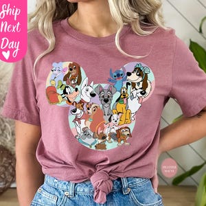 May include: Mauve-colored t-shirt featuring a graphic of various cartoon dogs arranged in a Mickey Mouse head shape. The shirt has short sleeves and a crew neck. The text "Ship Next Day" is on the upper left side of the shirt.