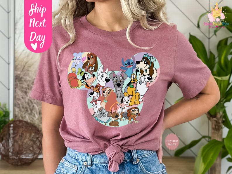 May include: Mauve-colored t-shirt featuring a graphic of various cartoon dogs arranged in a Mickey Mouse head shape. The shirt has short sleeves and a crew neck. The text "Ship Next Day" is on the upper left side of the shirt.