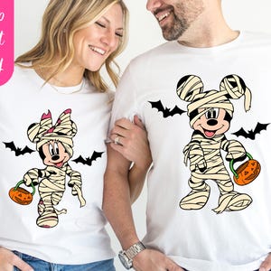 May include: White t-shirts featuring Mickey and Minnie Mouse dressed as mummies for Halloween. Each character holds a jack-o'-lantern and is surrounded by black bat silhouettes. The shirts have the text "White" and "Ship Next Day".