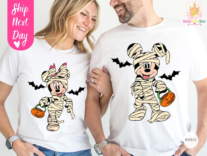 May include: White t-shirts featuring Mickey and Minnie Mouse dressed as mummies for Halloween. Each character holds a jack-o'-lantern and is surrounded by black bat silhouettes. The shirts have the text "White" and "Ship Next Day".