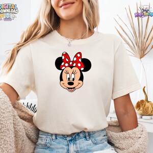以下が含まれることがあります： 中央にミニーマウスのグラフィックがプリントされたクリーム色の半袖Tシャツ。キャラクターは特徴的な黒い耳と、白い水玉模様が入った鮮やかな赤色のリボンを身につけており、明るい色の生地にデザインが配置されています。
