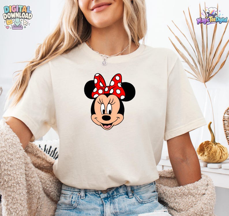 以下が含まれることがあります： 中央にミニーマウスのグラフィックがプリントされたクリーム色の半袖Tシャツ。キャラクターは特徴的な黒い耳と、白い水玉模様が入った鮮やかな赤色のリボンを身につけており、明るい色の生地にデザインが配置されています。