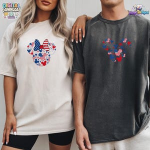 Puede incluir: Una camiseta de color crema con un gr&aacute;fico de una silueta de cabeza de rat&oacute;n compuesta por estrellas y rayas rojas, blancas y azules, y una camiseta gris carb&oacute;n oscuro con un gr&aacute;fico similar de cabeza de rat&oacute;n en patrones rojos, blancos y azules.
