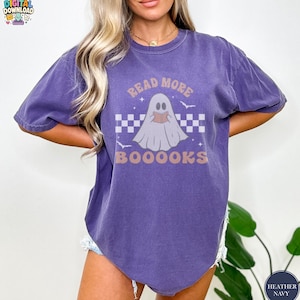 Op de afbeelding: Een heather navy t-shirt met korte mouwen en een afbeelding van een witte geest die een boek leest. Het ontwerp bevat een ruitpatroon, kleine vleermuizen en de tekst "READ MORE BOOOOKS" in een lichtbruine kleur. Het shirt heeft een ronde hals.