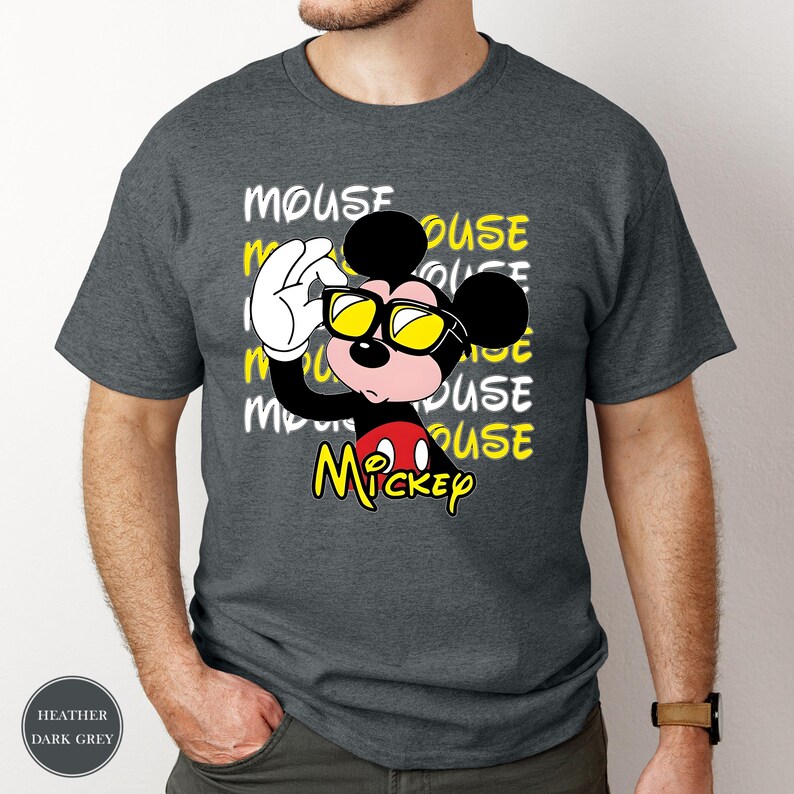 May include: Heather dark grey t-shirt featuring a graphic of Mickey Mouse wearing sunglasses. The word "MOUSE" is repeated in white and yellow. The word "Mickey" is written in yellow. The shirt is a crew neck.