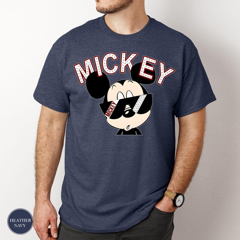 May include: Heather navy blue t-shirt featuring a cartoon Mickey Mouse wearing sunglasses. The word "MICKEY" is arched above the character in white letters with red accents. The shirt is a classic crew neck style.