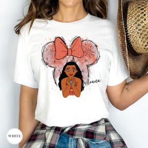 May include: White t-shirt featuring a graphic of Moana with a Minnie Mouse-inspired bow and ears. The design includes the word "Moana" in a script font. The shirt has a crew neck and short sleeves.