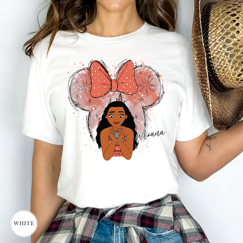 May include: White t-shirt featuring a graphic of Moana with a Minnie Mouse-inspired bow and ears. The design includes the word "Moana" in a script font. The shirt has a crew neck and short sleeves.