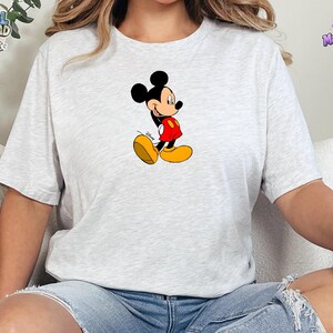 Peut inclure: Un t-shirt &agrave; manches courtes gris chin&eacute; clair avec un motif central repr&eacute;sentant Mickey Mouse. Le personnage est illustr&eacute; dans une pose classique, portant un short rouge et de grandes chaussures jaunes, avec le logo Disney en noir.