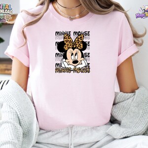Pode incluir: Uma t-shirt de manga curta rosa claro com um gr&aacute;fico da Minnie Mouse usando um la&ccedil;o com estampa de leopardo. O design inclui o texto Minnie Mouse repetido em fonte de contorno preto atr&aacute;s da personagem e na parte inferior em estampa de leopardo.