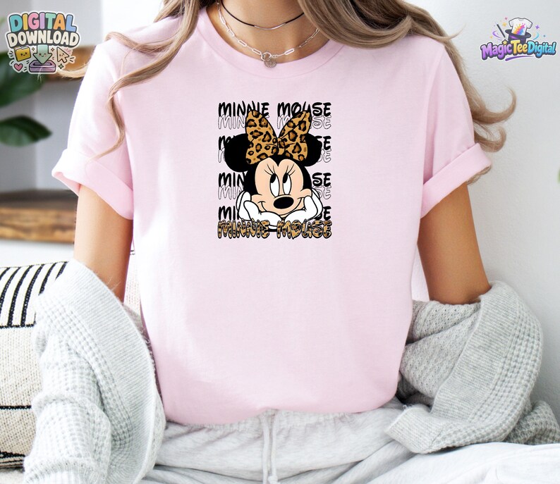 Pode incluir: Uma t-shirt de manga curta rosa claro com um gr&aacute;fico da Minnie Mouse usando um la&ccedil;o com estampa de leopardo. O design inclui o texto Minnie Mouse repetido em fonte de contorno preto atr&aacute;s da personagem e na parte inferior em estampa de leopardo.