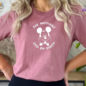 Puede incluir: Una camiseta de manga corta de color malva con un gr&aacute;fico blanco de Mickey Mouse y el texto "THE HAPPIEST DAD ON EARTH" impreso en disposici&oacute;n circular alrededor del personaje. La camiseta tiene cuello redondo y corte est&aacute;ndar.