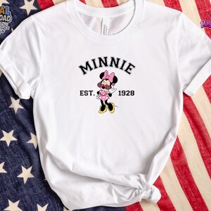 May include: A white short-sleeved t-shirt featuring a graphic of Minnie Mouse wearing pink sunglasses and a polka dot dress. The text above the character reads MINNIE in a black arched font, with EST. 1928 printed in black on either side of the figure.