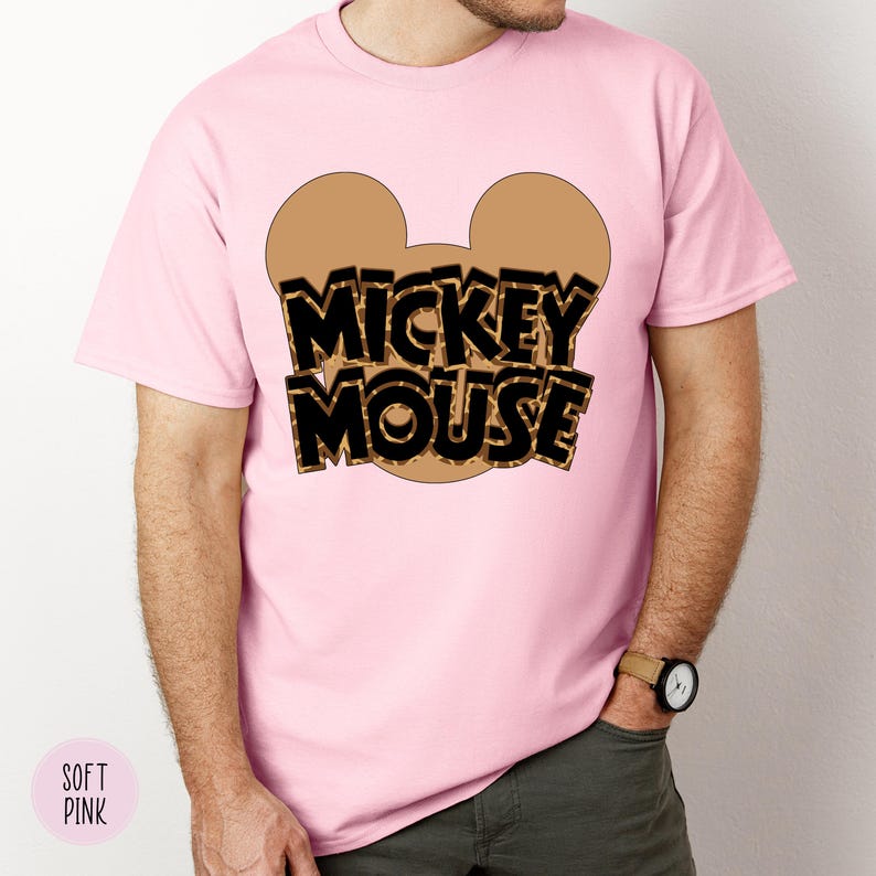 Puede incluir: Una camiseta rosa claro con un contorno de cabeza de Mickey Mouse marr&oacute;n y las palabras "MICKEY MOUSE" en estampado de leopardo negro y marr&oacute;n. La camiseta es de cuello redondo y la lleva una persona.