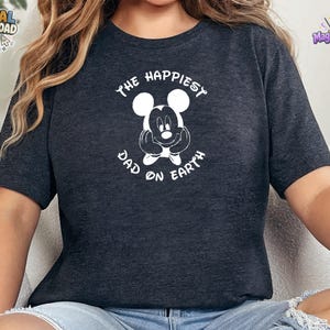 Puede incluir: Una camiseta de manga corta en gris jaspeado oscuro con un gr&aacute;fico blanco de una cabeza de rat&oacute;n de dibujos animados con las manos apoyadas en las mejillas. El texto "THE HAPPIEST DAD ON EARTH" est&aacute; impreso en blanco en arco.