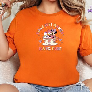 May include: An orange t-shirt featuring a graphic print of Daisy Duck and Minnie Mouse sitting in a teacup. The text "GIRLS JUST WANNA HAVE FUN!" is printed in a curved, colorful font above and below the characters, surrounded by small hearts.