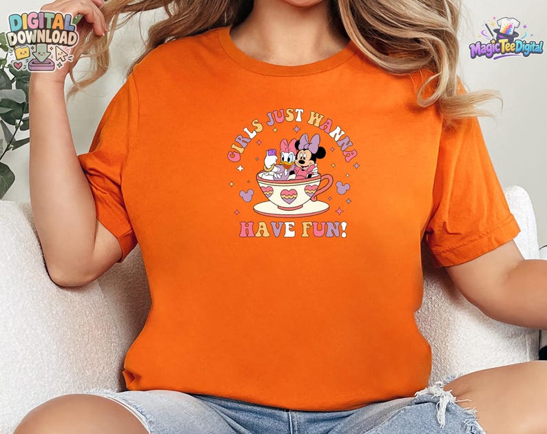 May include: An orange t-shirt featuring a graphic print of Daisy Duck and Minnie Mouse sitting in a teacup. The text "GIRLS JUST WANNA HAVE FUN!" is printed in a curved, colorful font above and below the characters, surrounded by small hearts.