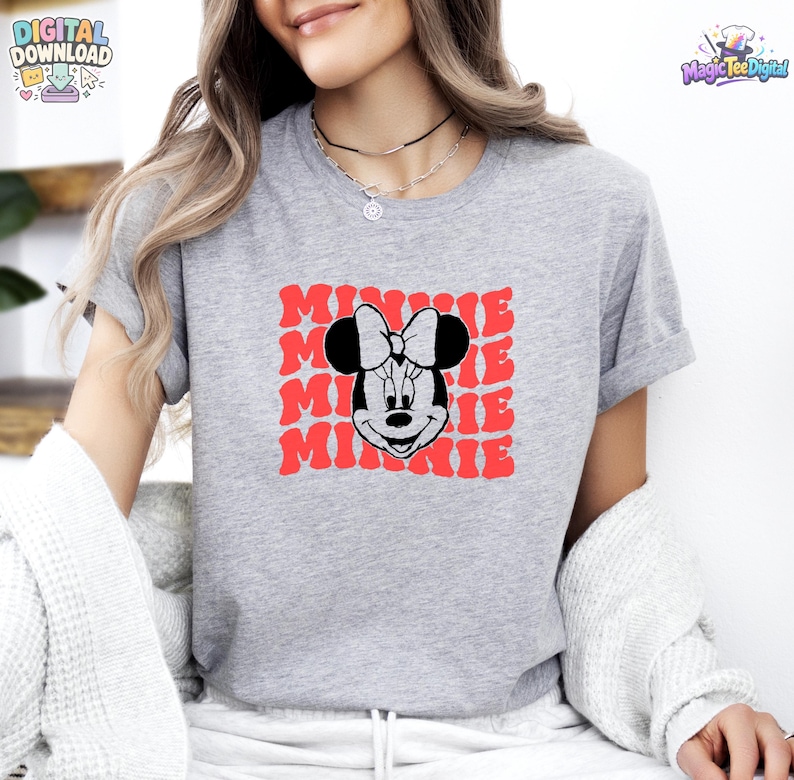 May include: A heather gray short-sleeve t-shirt featuring a black and white graphic of Minnie Mouse centered on the chest. Behind the character, the word MINNIE is repeated in a wavy, red, retro-style font. The shirt has a crew neckline.