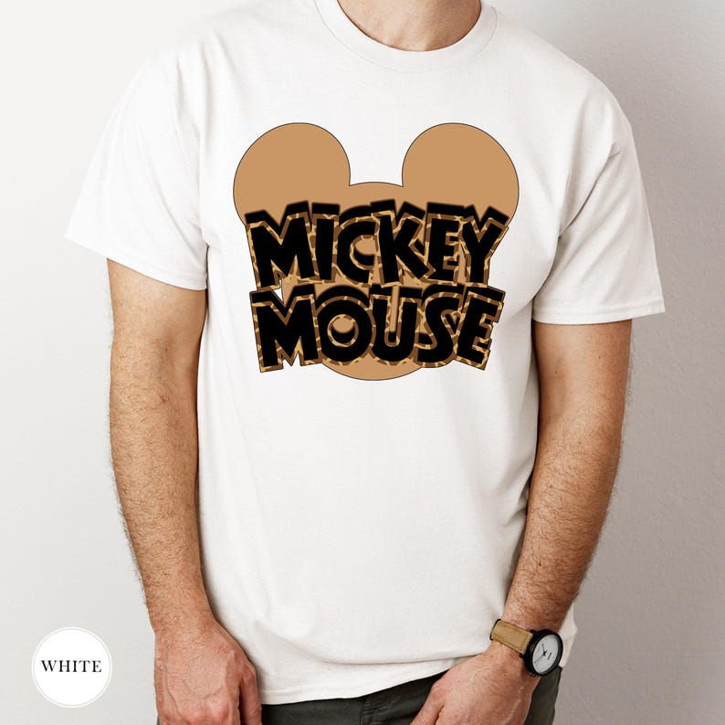 Puede incluir: Camiseta blanca con un contorno de cabeza de Mickey Mouse marr&oacute;n y las palabras "MICKEY MOUSE" en negro y marr&oacute;n. La camiseta es de cuello redondo.