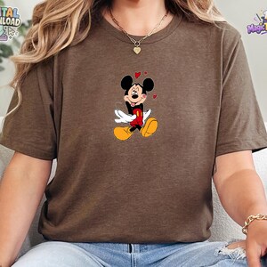 May include: A brown short-sleeved t-shirt featuring a graphic of Mickey Mouse with his tongue sticking out, wearing red shorts and yellow shoes, surrounded by small red hearts. The design is centered on the chest of the crew neck garment.