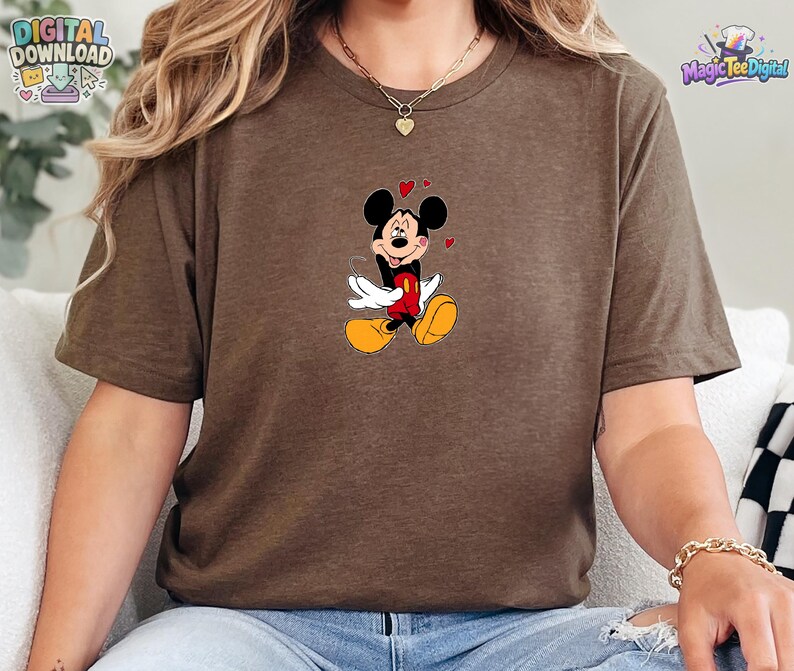 May include: A brown short-sleeved t-shirt featuring a graphic of Mickey Mouse with his tongue sticking out, wearing red shorts and yellow shoes, surrounded by small red hearts. The design is centered on the chest of the crew neck garment.
