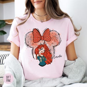 Puede incluir: Camiseta rosa claro con una ilustraci&oacute;n de dibujos animados de Merida de la pel&iacute;cula de Disney Brave. El dise&ntilde;o incluye un lazo rojo con lunares blancos, un personaje de Merida y el nombre "Merida" en escritura.