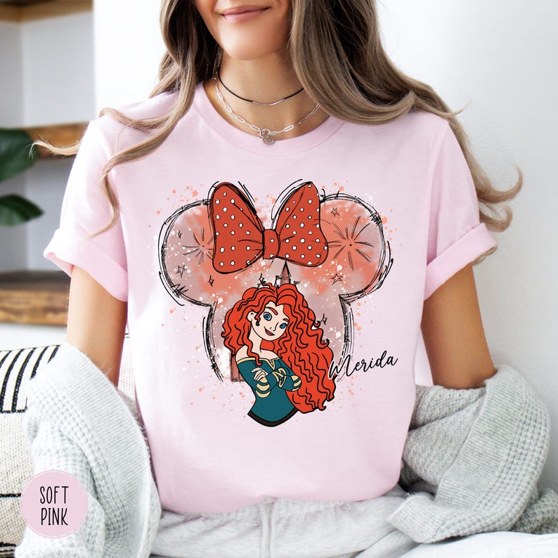 Puede incluir: Camiseta rosa claro con una ilustraci&oacute;n de dibujos animados de Merida de la pel&iacute;cula de Disney Brave. El dise&ntilde;o incluye un lazo rojo con lunares blancos, un personaje de Merida y el nombre "Merida" en escritura.