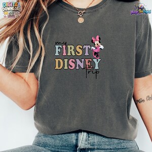 May include: A charcoal grey short-sleeve t-shirt featuring a graphic print with the text "my FIRST DISNEY TRIP" in multi-coloured pastel letters. A small illustration of Minnie Mouse in a pink dress is positioned to the right of the text.