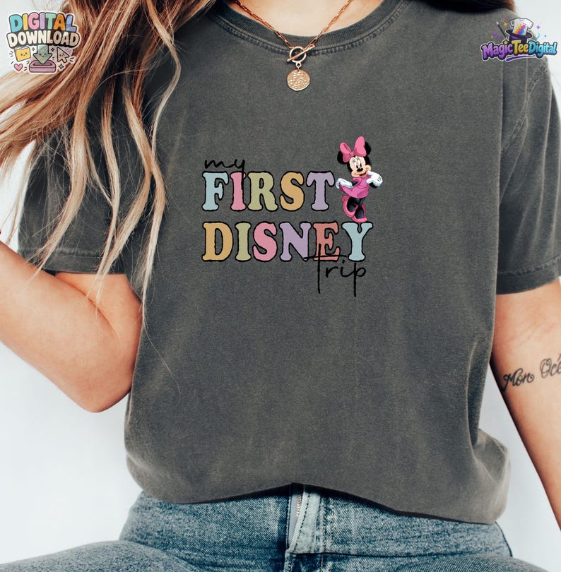 May include: A charcoal grey short-sleeve t-shirt featuring a graphic print with the text "my FIRST DISNEY TRIP" in multi-coloured pastel letters. A small illustration of Minnie Mouse in a pink dress is positioned to the right of the text.