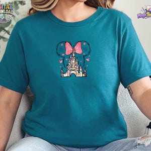 Pu&ograve; includere: Una t-shirt color verde acqua a maniche corte con una grafica centrale che raffigura la sagoma di un castello sormontata da un fiocco rosa a pois. Il design include contorni neri abbozzati di orecchie di topo, piccoli cuori rosa e stelle.
